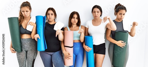 Group of women holding yoga mat standing over isolated background pointing down looking sad and upset, indicating direction with fingers, unhappy and depressed.