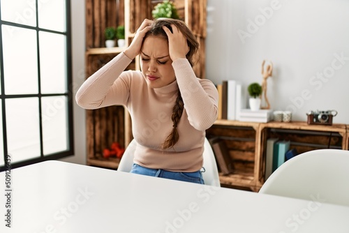 Young hispanic girl wearing casual clothes sitting on the table at home suffering from headache desperate and stressed because pain and migraine. hands on head.