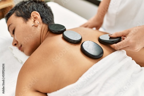 Young hispanic woman having back massage using black hot stones at beauty center