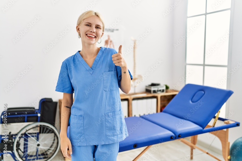 Beautiful caucasian physiotherapist woman working at pain recovery clinic doing happy thumbs up gesture with hand. approving expression looking at the camera showing success. Beautiful caucasian physiotherapist woman working at pain recovery clinic doing happy thumbs up gesture with hand. approving expression looking at the camera showing success.