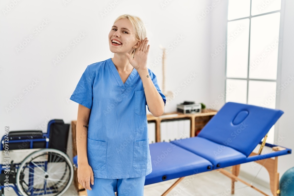 Beautiful caucasian physiotherapist woman working at pain recovery clinic smiling with hand over ear listening an hearing to rumor or gossip. deafness concept. Beautiful caucasian physiotherapist woman working at pain recovery clinic smiling with hand over ear listening an hearing to rumor or gossip. deafness concept.