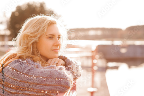 Foto Close-up of young woman with long wavy fair hair wearing brown woolen cardigan, standing at concrete river embankment