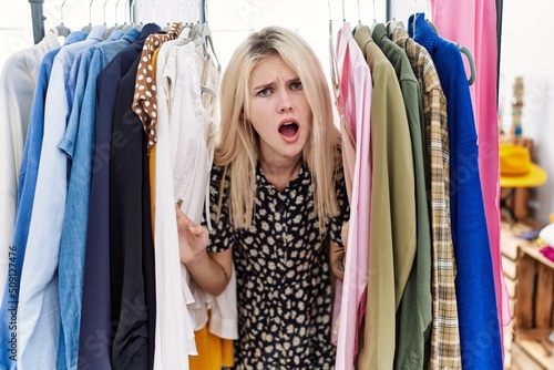 Young blonde woman searching clothes on clothing rack in shock face, looking skeptical and sarcastic, surprised with open mouth