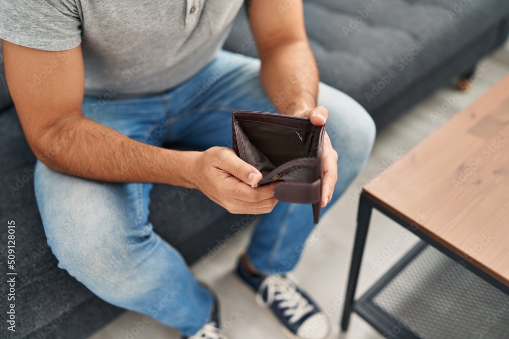 Young hispanic man showing empty wallet sitting on sofa at home Young hispanic man showing empty wallet sitting on sofa at home