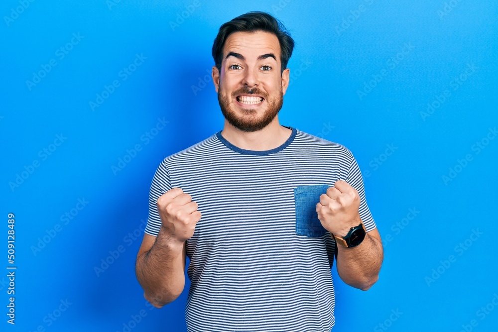 Handsome caucasian man with beard wearing casual striped t shirt celebrating surprised and amazed for success with arms raised and open eyes. winner concept. Handsome caucasian man with beard wearing casual striped t shirt celebrating surprised and amazed for success with arms raised and open eyes. winner concept.