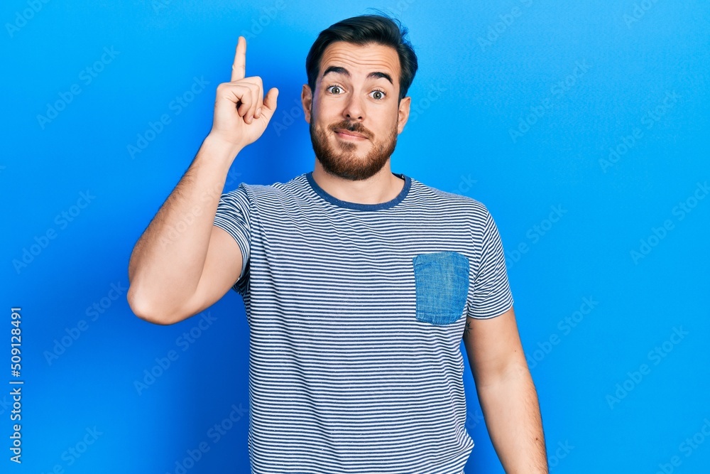 Fototapeta premium Handsome caucasian man with beard wearing casual striped t shirt pointing finger up with successful idea. exited and happy. number one.