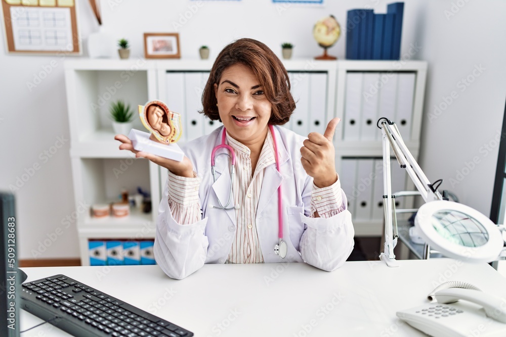 Middle age hispanic doctor woman holding anatomical model of female uterus with fetus pointing thumb up to the side smiling happy with open mouth Middle age hispanic doctor woman holding anatomical model of female uterus with fetus pointing thumb up to the side smiling happy with open mouth