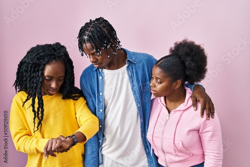 Group of three young black people standing together over pink background checking the time on wrist watch, relaxed and confident
