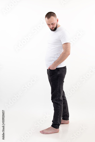 Full-length sad and upset barefoot bearded man in white t-shirt, jeans posing, look down. Fast gain weight. Not sporty