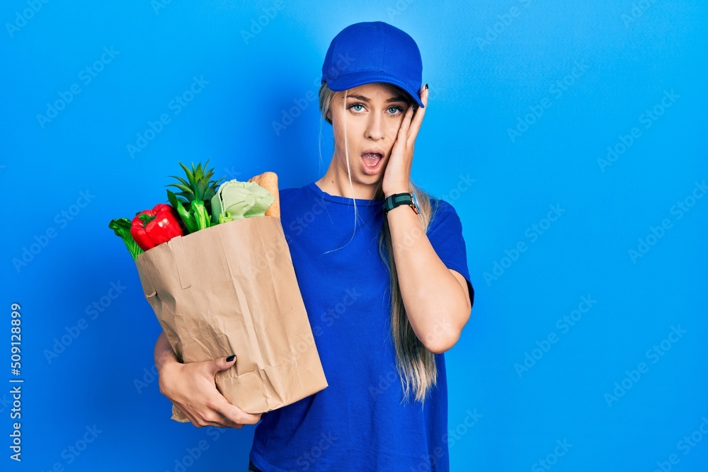 Young caucasian woman wearing courier uniform with groceries from supermarket yawning tired covering half face, eye and mouth with hand. face hurts in pain. Young caucasian woman wearing courier uniform with groceries from supermarket yawning tired covering half face, eye and mouth with hand. face hurts in pain.