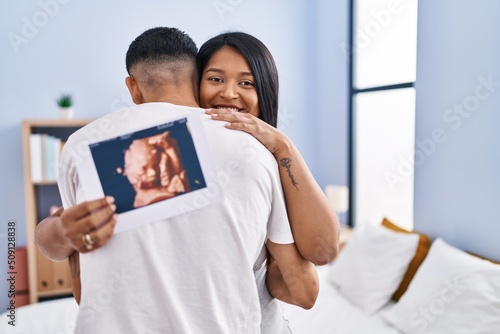Young latin couple expecting baby hugging each other holding ecography at bedroom photo