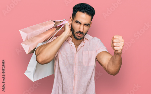 Foto Hispanic man with beard holding shopping bags annoyed and frustrated shouting wi