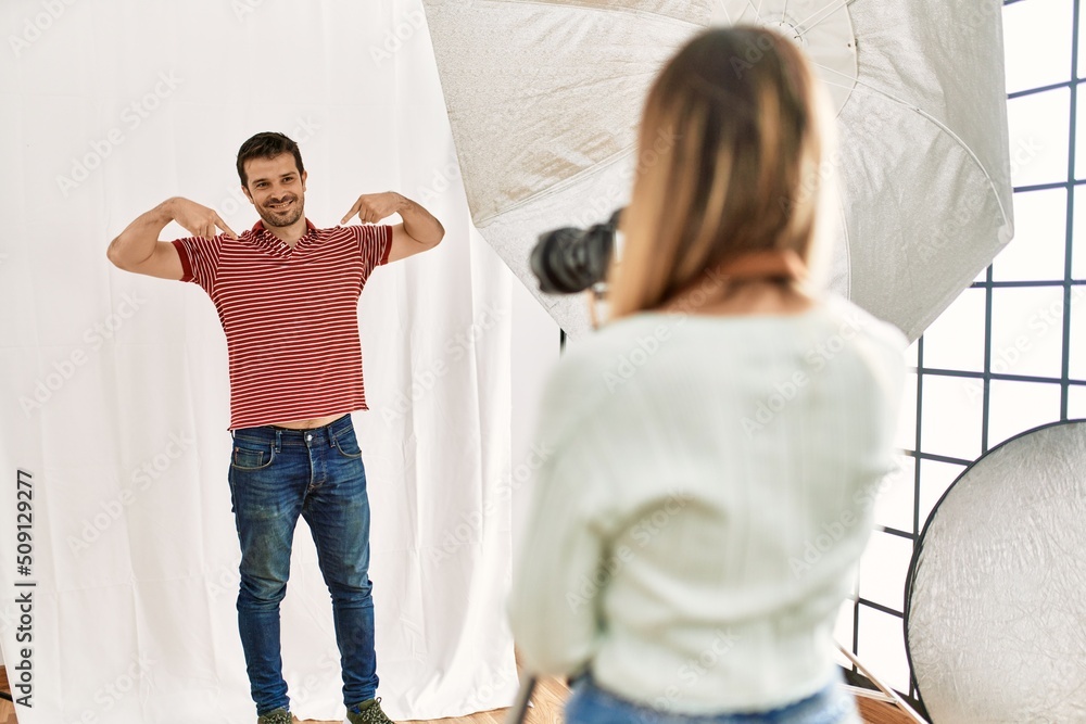 Fototapeta premium Woman photographer talking pictures of man posing as model at photography studio looking confident with smile on face, pointing oneself with fingers proud and happy.