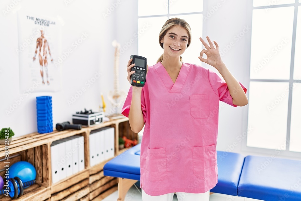 Young physiotherapist woman working at pain recovery clinic holding dataphone doing ok sign with fingers, smiling friendly gesturing excellent symbol Young physiotherapist woman working at pain recovery clinic holding dataphone doing ok sign with fingers, smiling friendly gesturing excellent symbol