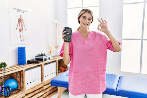 Young physiotherapist woman working at pain recovery clinic holding dataphone doing ok sign with fingers  smiling friendly gesturing excellent symbol