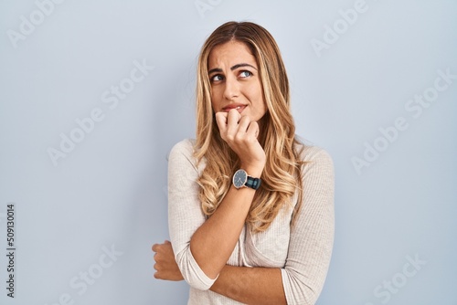 Young blonde woman standing over isolated background looking stressed and nervous with hands on mouth biting nails. anxiety problem.