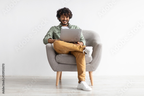 Joyful young indian sitting in armchair, chilling with modern laptop photo