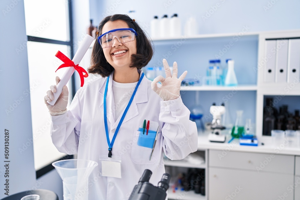 Young hispanic woman working at scientist laboratory holding degree doing ok sign with fingers, smiling friendly gesturing excellent symbol Young hispanic woman working at scientist laboratory holding degree doing ok sign with fingers, smiling friendly gesturing excellent symbol