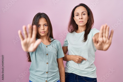 Young mother and daughter standing over pink background doing stop sing with palm of the hand. warning expression with negative and serious gesture on the face.