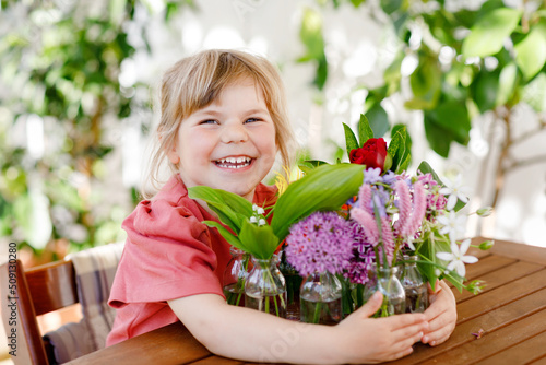 Little preschool girl with flower bouquet at home. Toddler child putting colorful garden summer flowers in small bottles with water. Home activities for kids. Flowers in rainbow colors. Happy child.