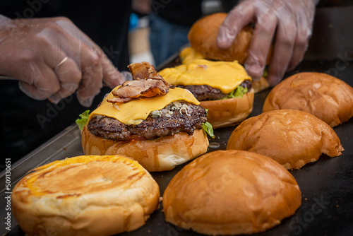Hands cooking or preparing burger or cheeseburger made of grilled beef meat, green lettuce, cheese and fried bacon between roasted bun bread served at barbeque street food festival at restaurant