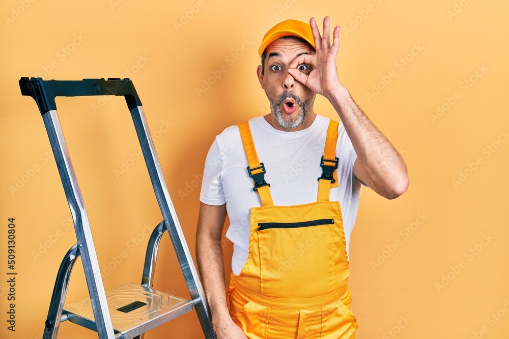 Handsome middle age man with grey hair holding ladder doing ok gesture shocked with surprised face, eye looking through fingers. unbelieving expression. Handsome middle age man with grey hair holding ladder doing ok gesture shocked with surprised face, eye looking through fingers. unbelieving expression.