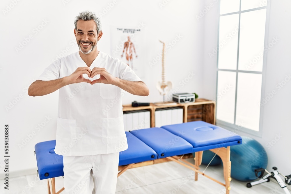 Middle age hispanic therapist man working at pain recovery clinic smiling in love doing heart symbol shape with hands. romantic concept.