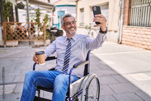 Middle age hispanic man sitting on wheelchair makes selfie by the smartphone at street