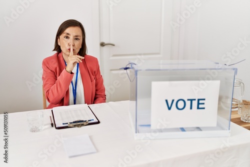 Beautiful middle age hispanic woman at political election sitting by ballot asking to be quiet with finger on lips. silence and secret concept.