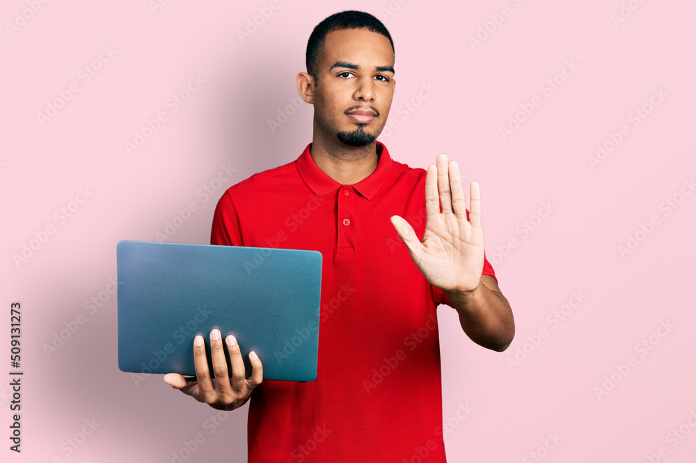 Young african american man working using computer laptop with open hand doing stop sign with serious and confident expression, defense gesture Young african american man working using computer laptop with open hand doing stop sign with serious and confident expression, defense gesture