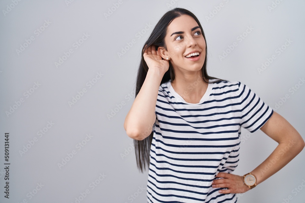 Young brunette woman wearing striped t shirt smiling with hand over ear listening an hearing to rumor or gossip. deafness concept. Young brunette woman wearing striped t shirt smiling with hand over ear listening an hearing to rumor or gossip. deafness concept.
