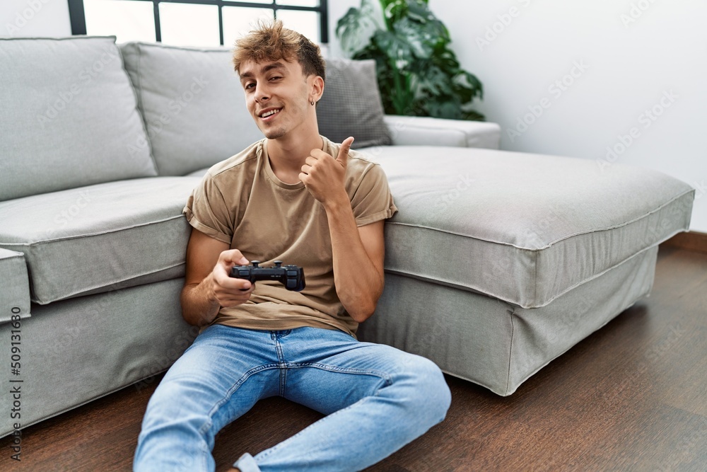Young caucasian man playing video game holding controller at home smiling happy and positive, thumb up doing excellent and approval sign