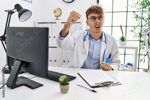 Young caucasian doctor man working at the clinic angry and mad raising fist frustrated and furious while shouting with anger. rage and aggressive concept.