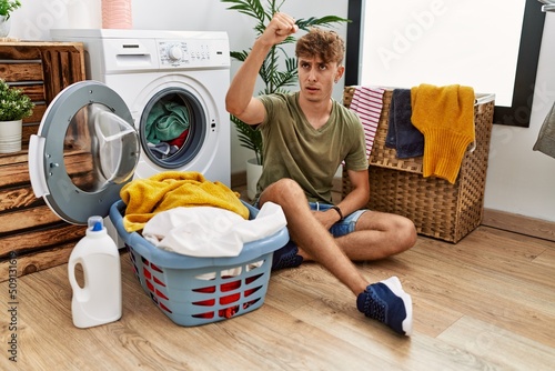 Young caucasian man putting dirty laundry into washing machine angry and mad raising fist frustrated and furious while shouting with anger. rage and aggressive concept.