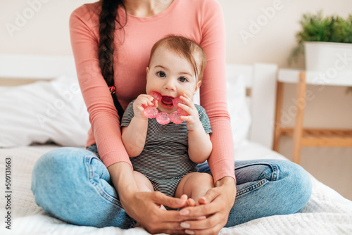 Cute little baby girl playing with teething toy, biting teether and looking at camera, sitting with mother on bed at home