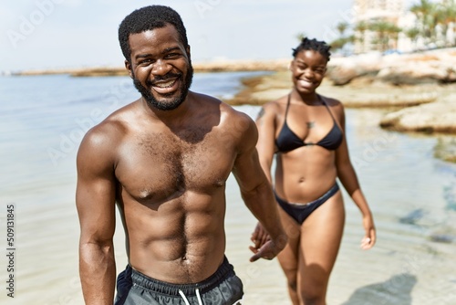 Young african american tourist couple wearing swimwear walking with hands together at the beach.