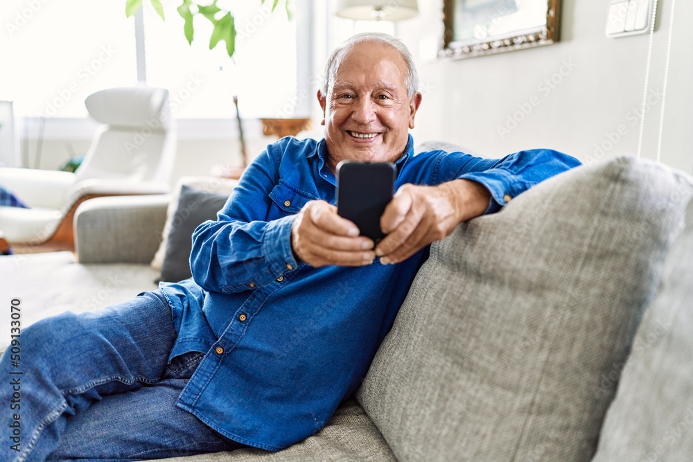 Senior man with grey hair sitting on the sofa at the living room of his house using smartphone