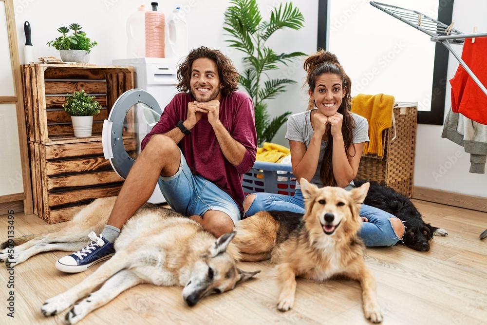 Young hispanic couple doing laundry with dogs laughing nervous and excited with hands on chin looking to the side