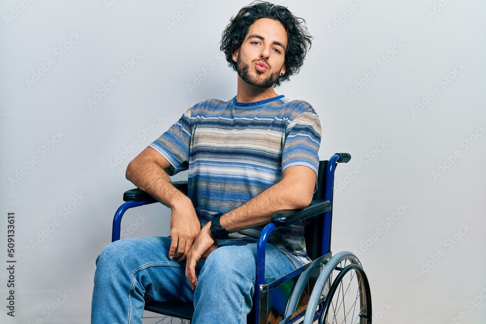 Handsome hispanic man sitting on wheelchair looking at the camera blowing a kiss on air being lovely and sexy. love expression. Handsome hispanic man sitting on wheelchair looking at the camera blowing a kiss on air being lovely and sexy. love expression.