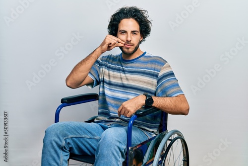 Handsome hispanic man sitting on wheelchair mouth and lips shut as zip with fingers. secret and silent, taboo talking