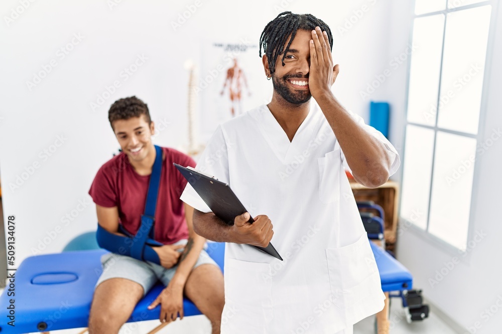 Obraz premium Young hispanic man working at pain recovery clinic with a man with broken arm covering one eye with hand, confident smile on face and surprise emotion.
