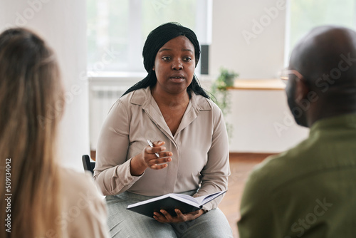 Young confident black woman with notebook and pen consulting intercultural couple sitting in front of her during session photo