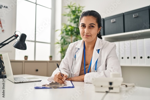 Young hispanic woman wearing doctor uniform writing medical report at clinic photo