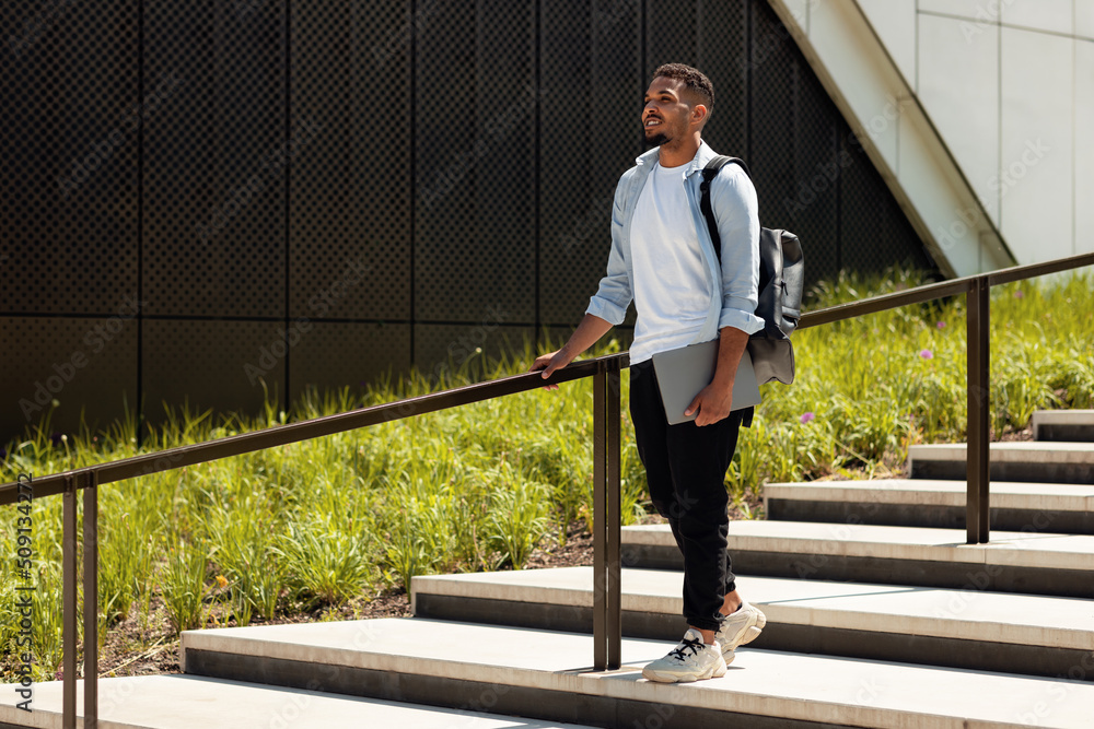Full length shot of african american man walking in park with laptop and backpack, resting after work or study outdoors