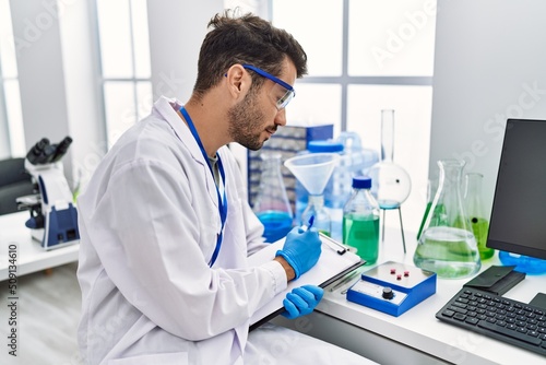 Young hispanic man wearing scientist uniform writing on clipboard working at laboratory