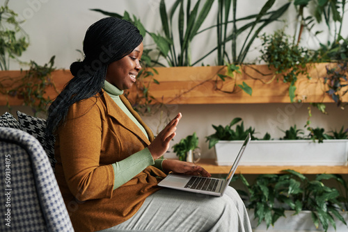 Side view of happy young psychotherapist with laptop on her knees waving hand to patient on screen during online session photo