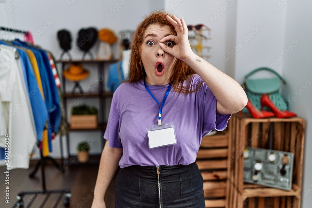 Young redhead woman working as manager at retail boutique doing ok gesture shocked with surprised face, eye looking through fingers. unbelieving expression.