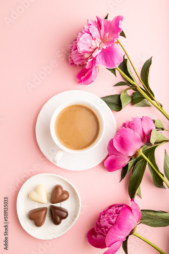 Cup of cioffee with chocolate candies, pink peony flowers on pink pastel background. top view, close up.