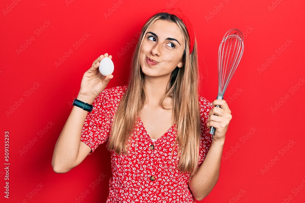 Beautiful hispanic woman holding egg and baker whisk smiling looking to the side and staring away thinking. Beautiful hispanic woman holding egg and baker whisk smiling looking to the side and staring away thinking.