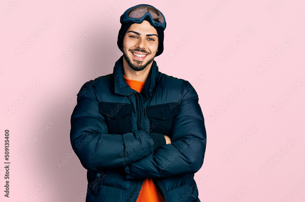 Young hispanic man with beard wearing snow wear and sky glasses happy face smiling with crossed arms looking at the camera. positive person. Young hispanic man with beard wearing snow wear and sky glasses happy face smiling with crossed arms looking at the camera. positive person.